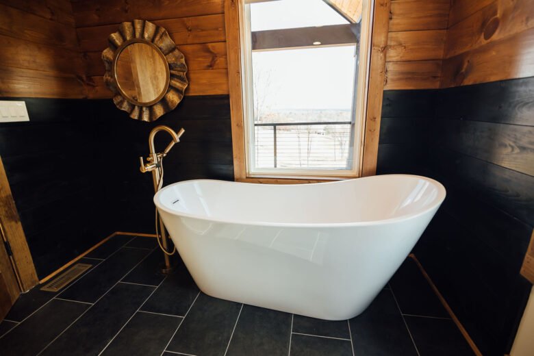 Freestanding white bathtub in a rustic bathroom with dark tile floors, wood walls, a window above the tub, and a decorative mirror on the wall.
