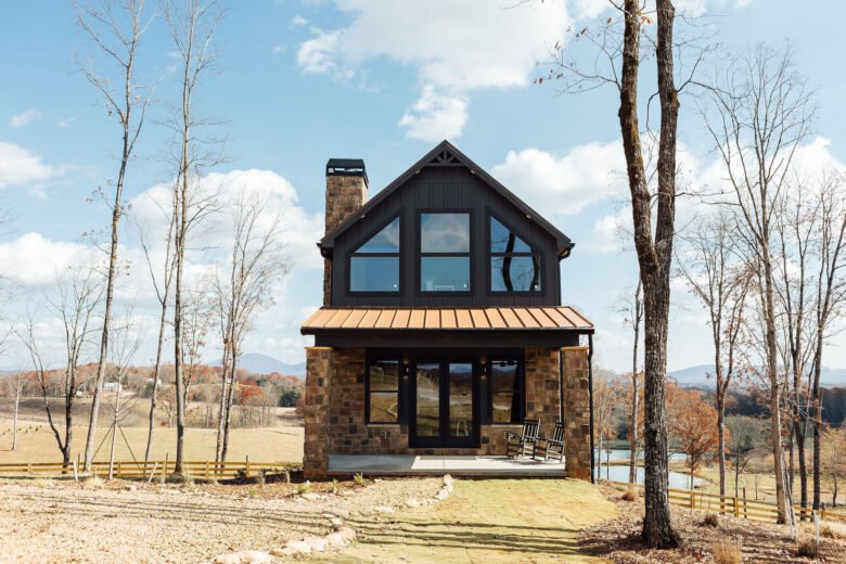 Two-story rustic cabin with large windows and a covered porch, set among leafless trees under a blue sky.