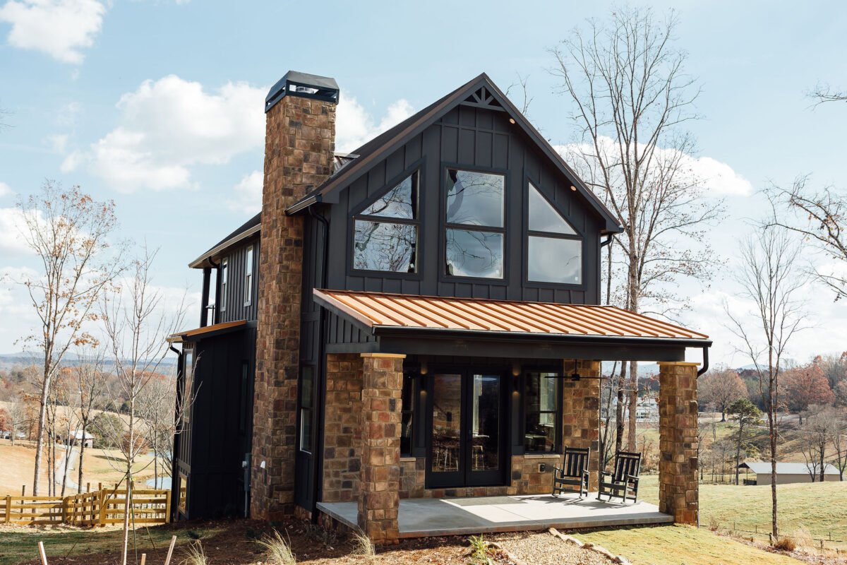 Two-story rustic cabin with a stone chimney, large front windows, and a covered porch, surrounded by trees and open lawn.