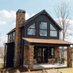Two-story rustic cabin with a stone chimney, large front windows, and a covered porch, surrounded by trees and open lawn.