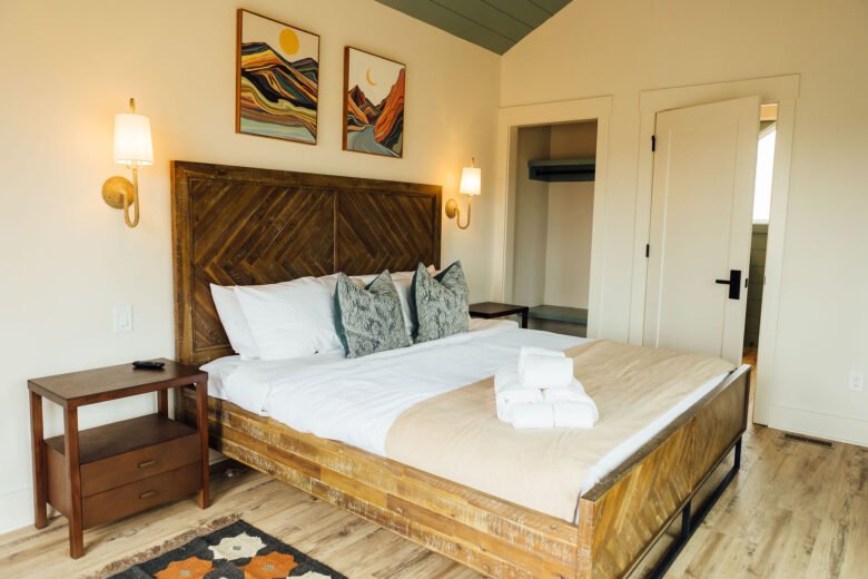 Rustic bedroom with a wood bed frame, white bedding and pillows, wall sconces, framed artwork above the headboard, and wood plank flooring.
