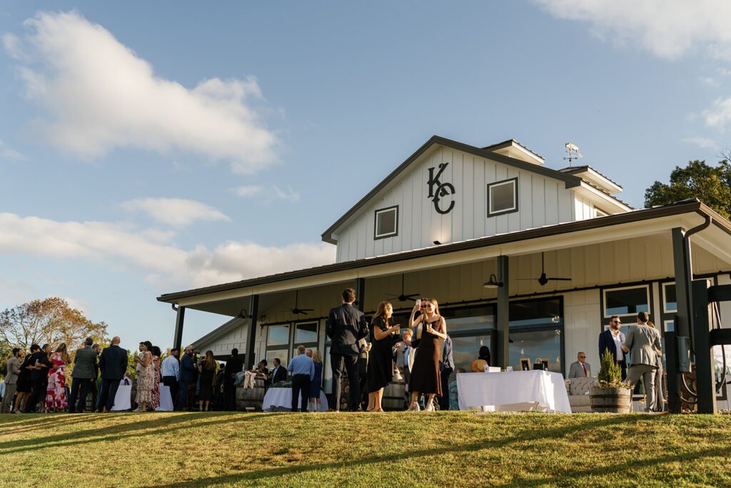 Guests mingling outdoors beside a rustic barn venue with a large covered patio and open lawn under a blue sky.