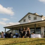 Guests mingling outdoors beside a rustic barn venue with a large covered patio and open lawn under a blue sky.