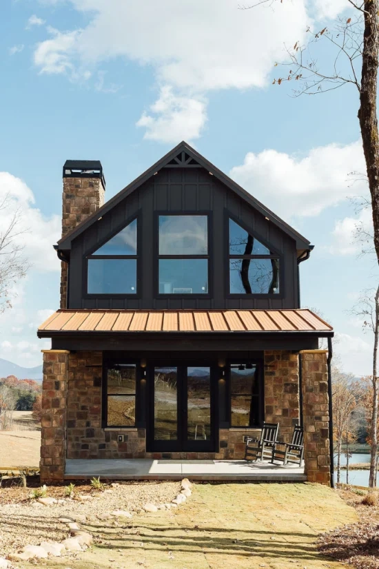 Dark modern lodge with a stone exterior, large windows, metal roof, front porch, and mountain views in the background.