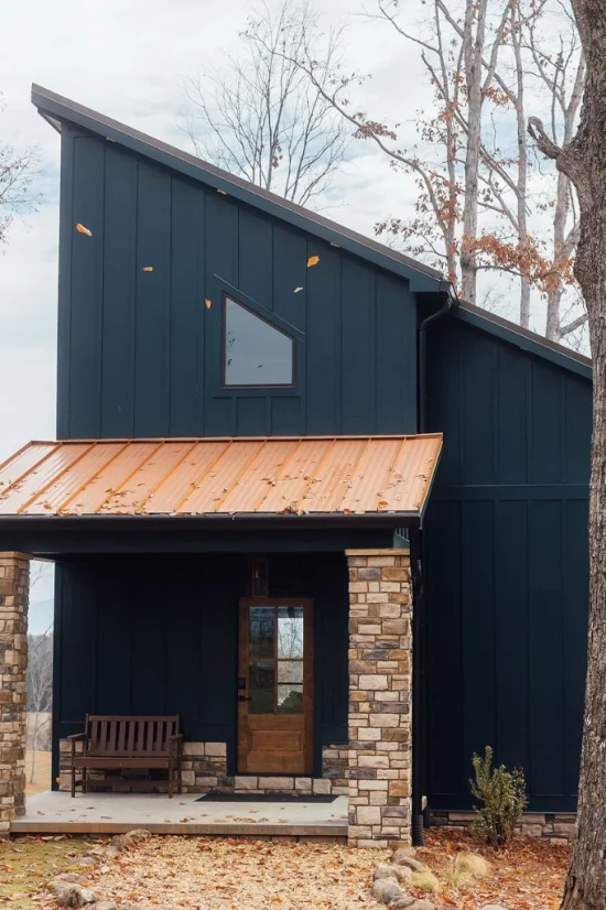 Modern dark lodge exterior with a sloped metal roof, stone columns, wooden front door, and covered porch surrounded by trees.