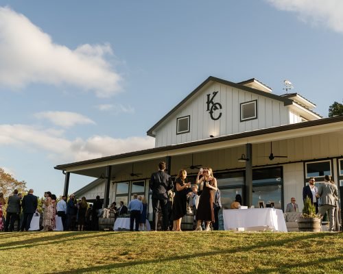 Guests mingling outdoors beside a rustic barn venue with a large covered patio and open lawn under a blue sky.